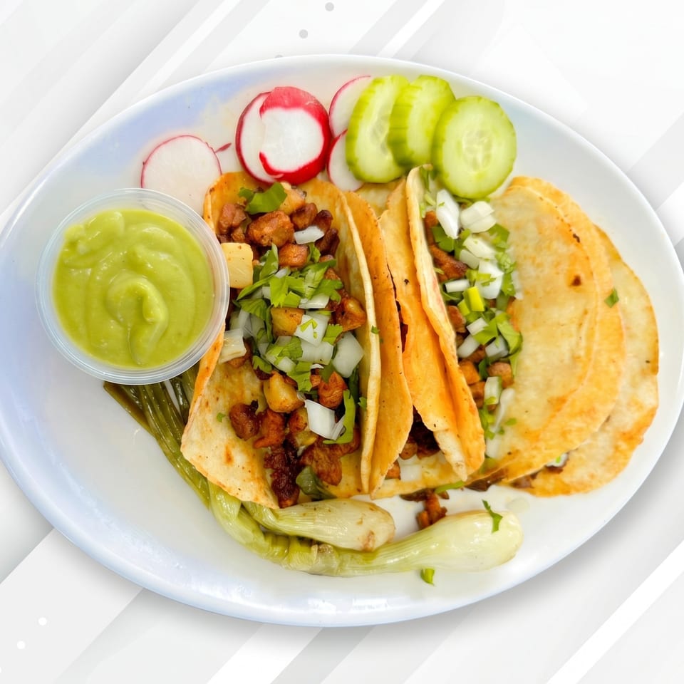 Three tacos with meat, onions, and cilantro, served with guacamole, radishes, cucumbers, and grilled green onions.